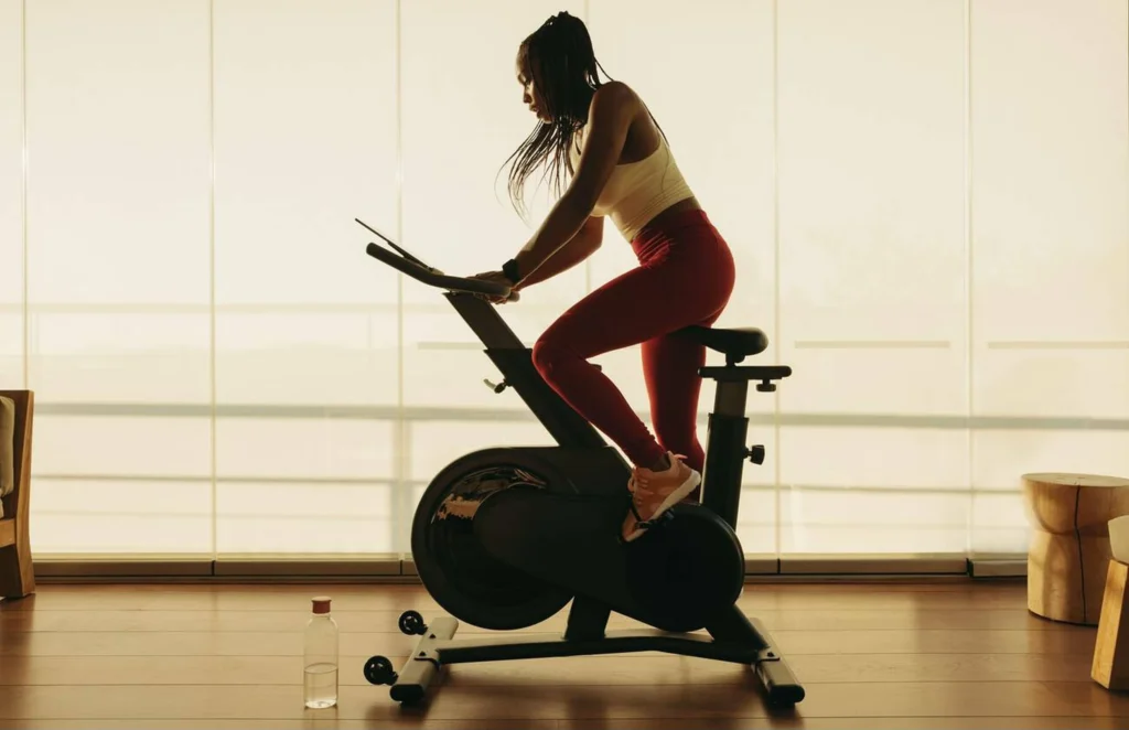 Mujer entrenando con monitor digital mientras realiza ciclo indoor para medir resistencia, calorías y ritmo cardíaco.