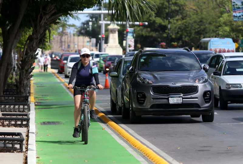 Mujer circulando en bicicleta junto a una fila de autos, sobre un carril exclusivo delimitado por bordes amarillos en una vía principal. Parte de la red de ciclopistas en Mérida